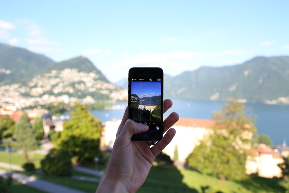 A person capturing a beautiful lake view with a smartphone, featuring mountains and buildings.