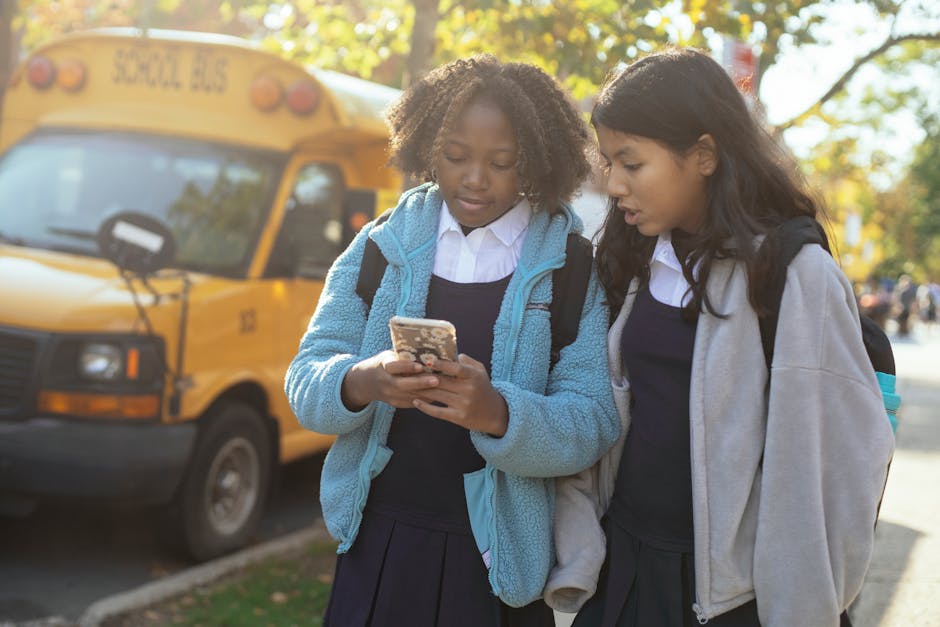 Two schoolgirls using a phone near a school bus, enjoying a sunny day, showcasing friendship and technology.