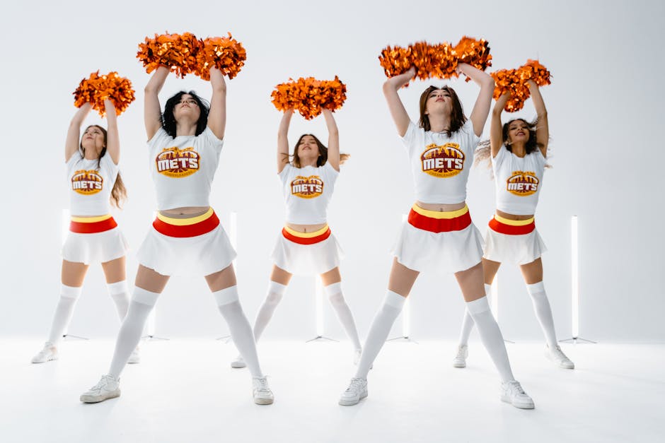 Five cheerleaders in coordinated uniforms performing a spirited dance routine indoors.