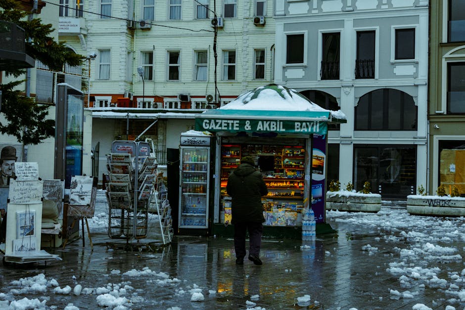 A man approaches a snow-covered newspaper kiosk on a wet winter day in the city.