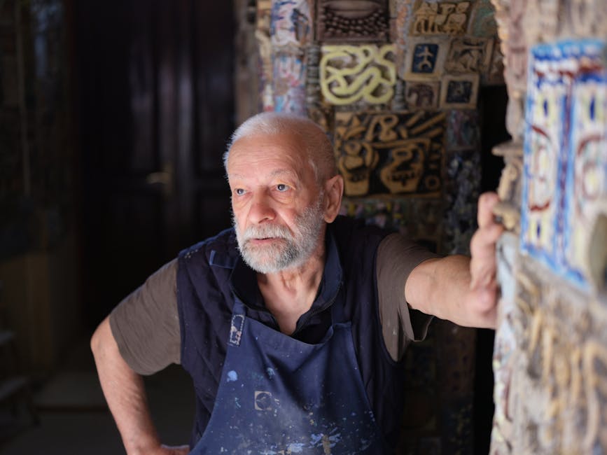 Elderly male artist wearing apron in a studio with artistic murals, thoughtfully gazing forward.