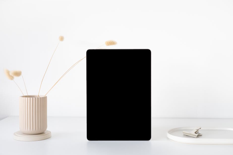 A minimalist mockup of an iPad on a white desk with a neutral vase and accessories, perfect for showcasing digital content.